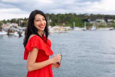 Deniz kenarında gülümseyen mutlu kadın. Kırmızı elbiseli neşeli bir kadın bulutlu bir günde elinde güneş gözlüğüyle marinanın yanında dururken ışıl ışıl gülümser.