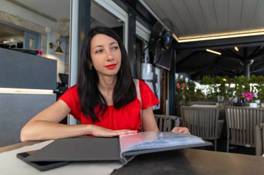 Kırmızı elbiseli kadın restoran menüsünden seçim yapıyor. Kırmızı elbiseli düşünceli bir kadın restoran masasında oturmuş, rahat bir yemek atmosferinin tadını çıkarırken menüyü karıştırıyor.
