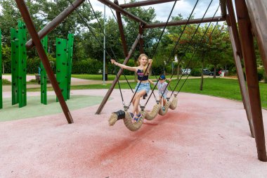 Çocuklar oyun parkında ip üzerinde salıncakta sallanıyor. Çağdaş bir oyun parkında salıncakta sallanan iki çocuk, yeşillik ve tırmanan yapılarla çevrili.