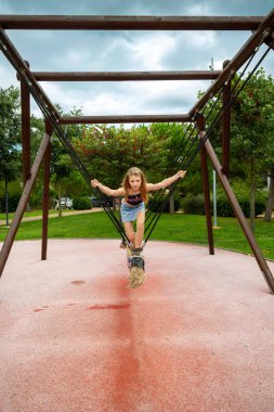 Kız çocuk parkında modern salıncakta dengede duruyor. Genç bir kız, ağaçlarla ve bulutlu gökyüzüyle çevrili parkta sallanan eşsiz bir ipte duruyor ve dengede duruyor.