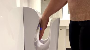 Kadın ellerini kurutuyor ve ellerini yaklaşan ellere tepki veren temas etmeyen bir kızılötesi sensör ile modern bir kurutucuda dezenfekte ediyor. Bir kadın ellerini otomatik el kurutma makinesinde kurutuyor. Duyusal el