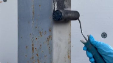 Açık havada çalışan bir sanatçı tarafından paten fırçasıyla metal bir sütun grisi boya. Boya silindirli eldivenli bir el. Dış onarım. Demir için özel korozyon önleyici boya. Paslı tedavinin