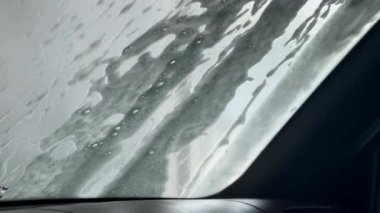 Suyun akışı arabanın ön camındaki köpüğü ve kiri temizler. Otomatik Oto Yıkama İşlemi. Araba camında sabun köpükleri, makro fotoğrafçılık. Beyaz sabun köpüğü araba camı ve dokusunda hareket eder.