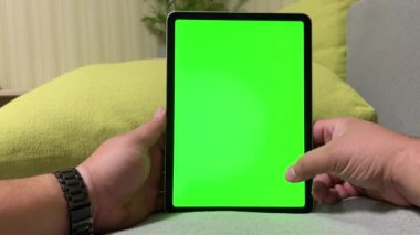 Bir adam elinde boş yeşil ekranlı dijital bir tablet tutuyor. Bir adam evde rahatlıyor, bilgisayar oyunları oynuyor ve web sitesine göz atıyor. Krom anahtar video şablonu. Krom anahtar - dikey ekran