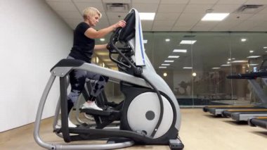 Orta yaşlı bir kadın spor için eliptik egzersiz makinesine biniyor. Tozluk ve beyaz spor ayakkabılı tombul bir kadın spor salonunda bir basamağa basar..