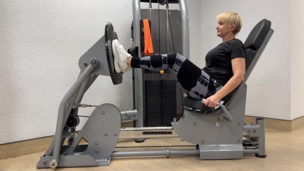 在体育馆里超重的55岁至60岁的女人 健身房里的一个女人在机器上压腿 一个成功女性过着健康生活的横向视频 — 图库视频影像