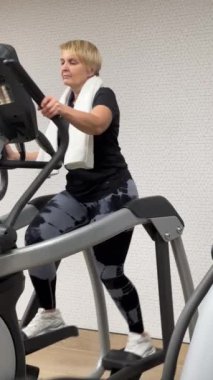 Spor salonundaki kilolu yaşlı bir kadın tüm kas grupları için kardiyo eğitimi alıyor. Yavaş çekim, dikey video. Sağlıklı yaşam tarzı kavramı.