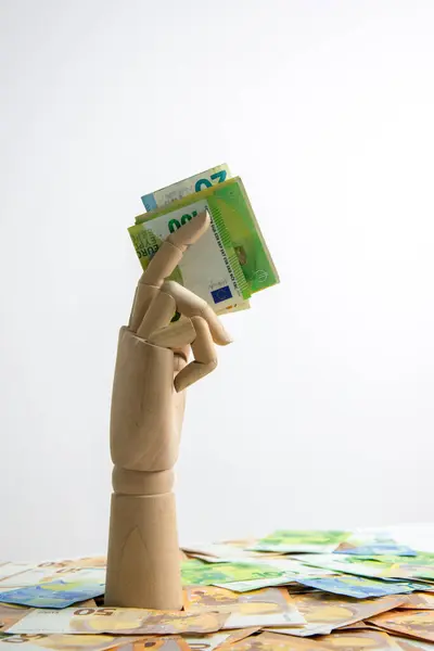 stock image A wooden hand reaches up from a pile of Euro banknotes, holding a few bills in its fingers.