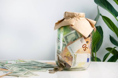 Etrafı dağınık banknotlar ve bozuk paralarla çevrili beyaz bir masanın üzerinde Avrupa ve ABD Doları banknotları olan bir kavanoz. Üzerinde kahverengi bir kese kağıdı ve arka planda yeşil yapraklar var..