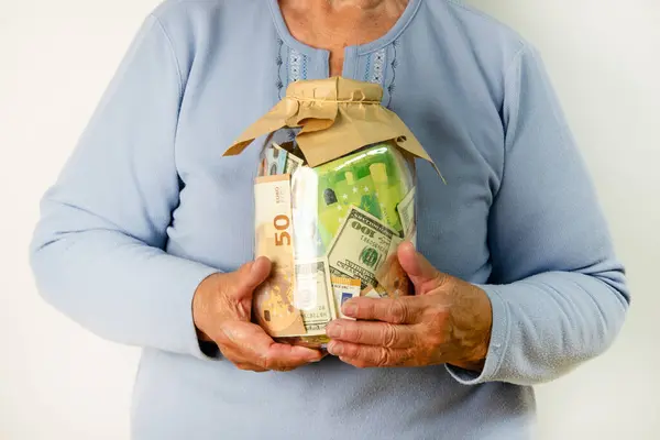 Yaşlı bir kadın, beyaz arka plan önünde dolar ve avro banknotlarının karışımıyla dolu cam bir kavanoz tutuyor..