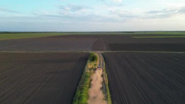 Bir dron geniş bir tarım arazisi üzerinde uçar, tarlaların arasından geçen toprak bir yol ortaya çıkarır..