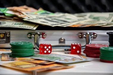 Kırmızı zarlar ve beyaz bir yüzeye yığılmış poker fişleri. Arka planda içi para dolu gümüş bir bavul görülüyor..