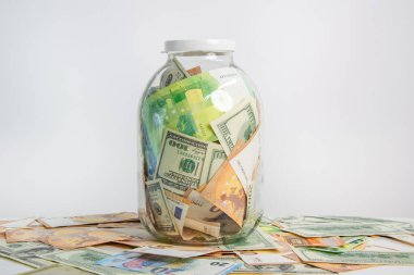 A glass jar filled with various US dollar and euro banknotes sits on a white surface, surrounded by scattered currency bills. clipart