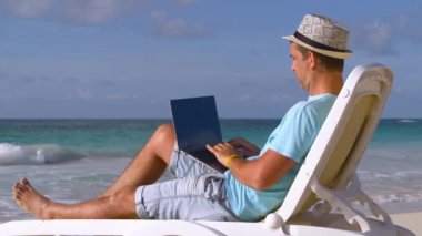 Plajda serbest çalışıyor. Güzel tropikal plajda serin serin otururken, yaz tatilinde serbest çalışan genç, beyaz bir adam dizüstü bilgisayarla çalışıyor. Yaz ve Tatil Konsepti