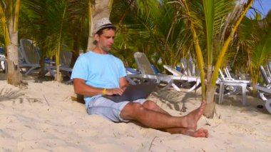 Serbest seyahat demek. Plajda dizüstü bilgisayarı olan serbest çalışan biri. İş ve tatil. Genç adam tropikal sahilde, palmiye ağaçlarının altında dizüstü bilgisayarda çalışıyor. Uzaktan kumandalı serbest çalışan bir adam. Çevrimiçi işler