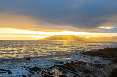 Deniz kenarındaki dağların üzerinde gün batımı. Dağlarda ve denizde dramatik bir gün batımı. Güneş ışınları bulutların arasından süzülüyor.