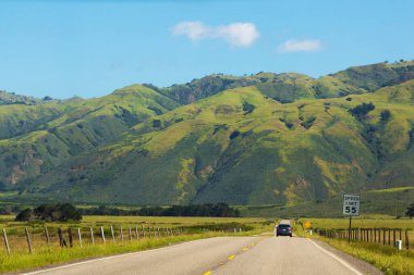 Yol, yeşil çayır ve dağların arka planına doğru uzanıyor. Bölünen şeritli asfalt yol mesafeye gidiyor. Otoyol boyunca araba sürüş, dikiz manzarası
