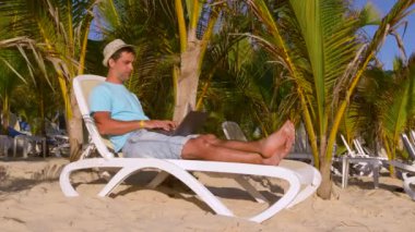 Laptop ve telefon kullanarak tropik bir plajda çalışan genç bir serbest çalışan. Yakışıklı genç adam bilgisayarına mesaj yazmaktan ve akıllı telefona bakmaktan mutlu. Genç işadamı tatilde.