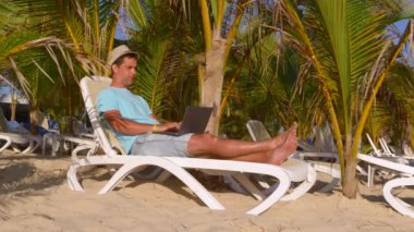 Sahilde güneşlenirken serbest çalışan biri. Serbest seyahat demek. İş ve tatil. Genç adam tropikal sahilde, palmiye ağaçlarının altında dizüstü bilgisayarda çalışıyor. Uzaktan kumandalı serbest çalışan bir adam. Çevrimiçi işler