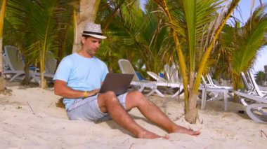 Genç serbest çalışan tropikal hedeften dizüstü bilgisayarla çalışıyor. Sahilde palmiye ağaçlarının altında oturan bir adam. Tropik palmiye ağaçları arasında dizüstü bilgisayarı olan serbest çalışan bir adam gün batımında adada çalışır..