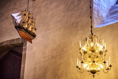 A model ship hangs from the ceiling in a Scandinavian church, a chandelier is lit, and a nautical emblem adorns the wall. The setting has a maritime theme, creating a cozy and historic atmosphere clipart