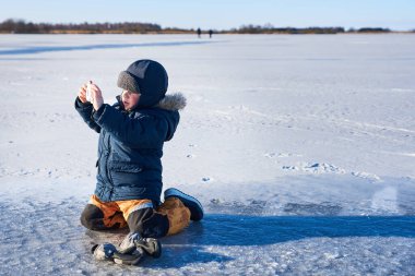 Bir çocuk donmuş bir gölün üzerinde diz çöker, gururla elinde bir oyuncak kamera tutar, kışlık kıyafetlerle sımsıkı sarılmıştır. Sahne, fotoğrafçılığın ve çocukluk macerasının huzurlu, karlı bir manzaradaki neşesini yakalıyor.