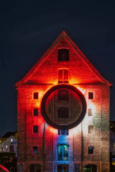 Bir tuğla bina, merkezinde asılı duran büyük siyah bir halkanın yer aldığı kırmızı, beyaz ve mavi ışıklarla çarpıcı bir şekilde aydınlatılır ve tarihi cepheye karşı çarpıcı ve modern bir kontrast oluşturur.