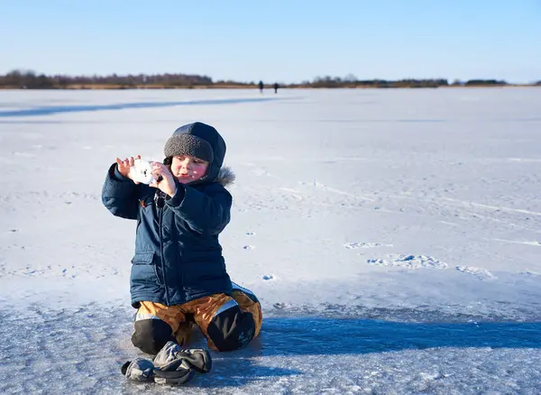 Kışlık giysili bir çocuk, elinde oyuncak bir kamerayla donmuş bir gölde diz çöker. Sahne, fotoğraf hobisinin ve çocukluk macerasının sakin, sakin kış manzarasındaki heyecanını yakalıyor.