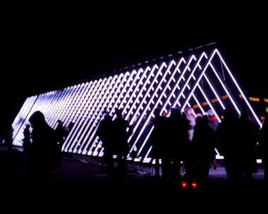 Siluetli figürler geceleri parıldayan bir üçgen ışık koridorunun önünden geçerek insan hareketini geometrik aydınlatmayla harmanlıyor. Teknoloji ve toplum arasındaki uyumun görsel metaforu