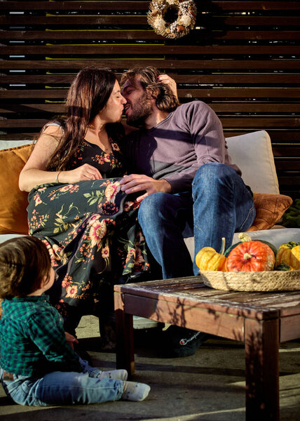 Couple (husband and wife) sits kissing on a rustic patio sofa, a child nearby framed by pumpkins and autumn vibe. The warm tones and seasonal decor evoke family intimacy, tradition and each other love