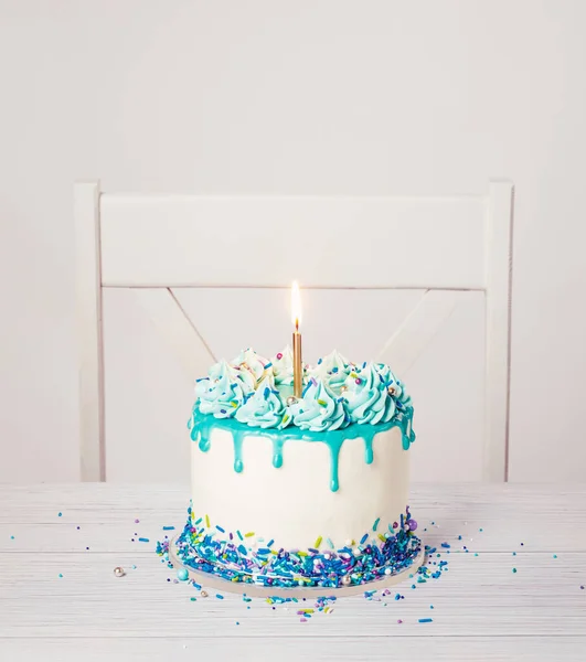 Blue and white buttercream birthday cake with a teal blue ganache drip, sprinkles, and bright gold candle on a white table and chair background. Trendy. Copy space.