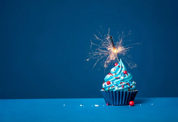 Festive buttercream birthday cupcake with red, white and blue sprinkles and lit sparkler on a dark blue background.