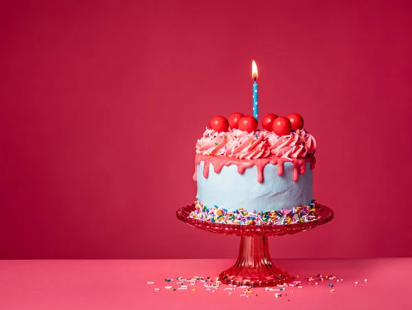 Bubble gum Birthday Drip Cake on a red stand with lit candle, colorful sprinkles, buttercream icing and red cherry candy balls on a vibrant pink background. Over the top fun. Copy space.