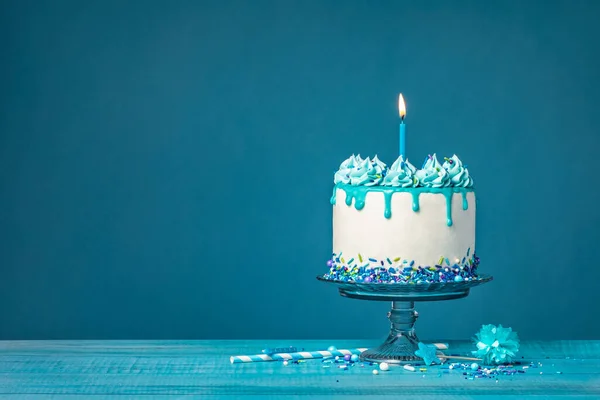 White drip cake with teal ganache, sprinkles, and a lit candle over a blue background. Simple and trendy. Copy space.