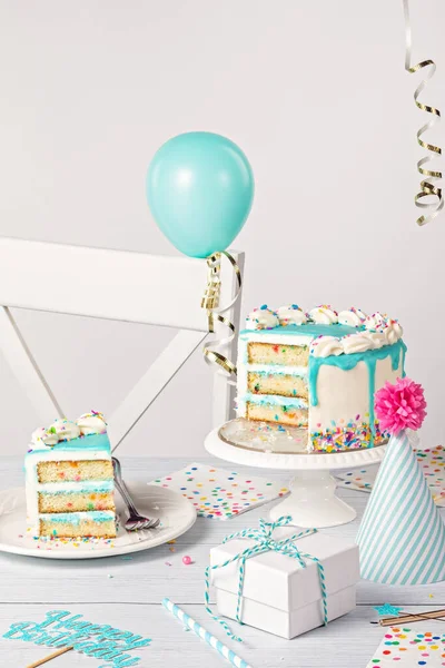 Happy Birthday party table setting with a sliced vanilla confetti cake, teal blue ganache drip, on light grey white background with balloons, ribbon decorations, hat and gifts.
