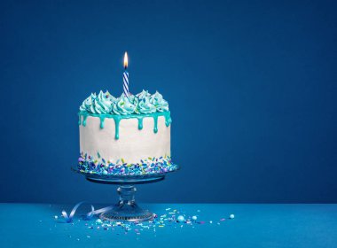 Beyaz doğum günü pastası, lacivert arka planda deniz mavisi ganache, serpiştirilmiş şeker ve yanan bir mum. Boşluğu kopyala.