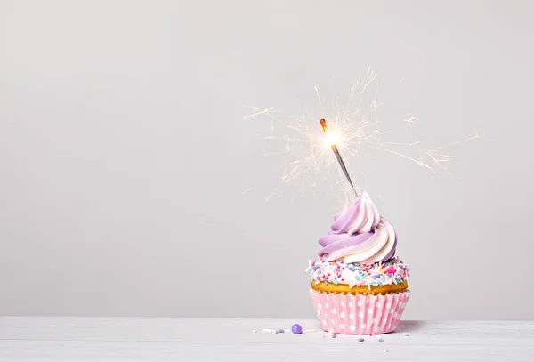 Purple and pink birthday celebration cupcake with buttercream icing swirl, sprinkles and lit sparkler on a light grey white background. copy space.