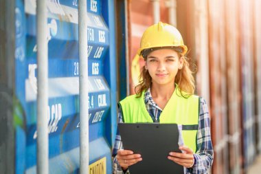 Kadın Mühendis, güneş batarken kargo konteynırı geçmişi olan PPE kontrol konteynırları kullanıyor. Lojistik küresel ithalat veya ihracat endüstriyel konsepti.