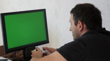 Young male gamer playing game console using joystick remote control over green screen monitor tv chroma key at home. Man Focused Gaming in Living Room