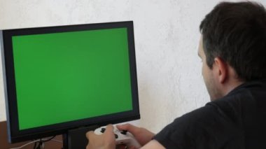 Young male gamer playing game console using joystick remote control over green screen monitor tv chroma key at home. Man Focused Gaming in Living Room