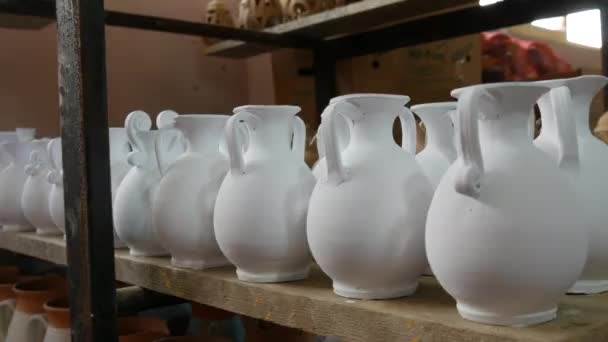 陶器工場のスタンドで作られた粘土や白いセラミック花瓶 ジャグや道具の多く オーブンからまだ絵付けされていないセラミック料理 — ストック動画