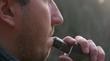 A handsome young man with a beard inhales the smoke of an electronic cigarette on the street close up view.