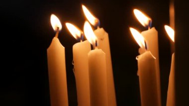 Long white religious candles, flames burning at night in the church. A womans hand lights and puts a candle to others.