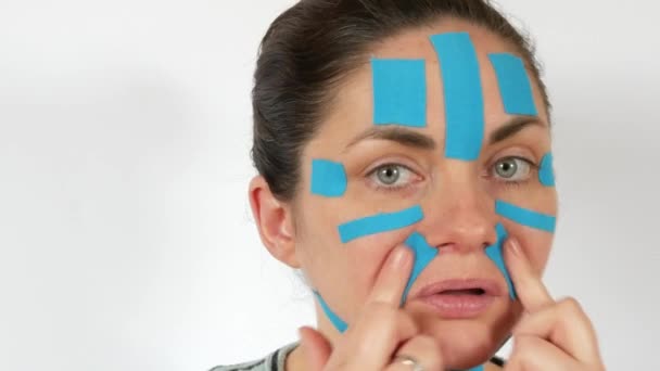 年轻女子用浅蓝色的动感条把脸 脖子贴在白色背景上 防止皮肤老化和皱纹 运动学和康复治疗 — 图库视频影像
