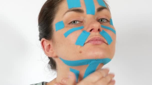 年轻女子用浅蓝色的动感条把脸 脖子贴在白色背景上 防止皮肤老化和皱纹 运动学和康复治疗 — 图库视频影像