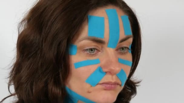 若い女性は白い背景に顔 首をテープライトブルーKinesioストリップを適用しました 肌の老化やしわと戦う 理学療法 運動療法と回復治療 — ストック動画