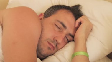Genç bir adam, elinde yeşil bir otel bileziği, beyaz bir yatak ve yastık üzerinde bir otel odasında uyuyor. Sağlıklı dinlenme ve uyku kavramı..