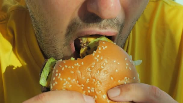 穿着黄色T恤的年轻人带着食欲吃汉堡包或汉堡包 嘴巴紧闭慢动作 咬和咀嚼垃圾食品 — 图库视频影像
