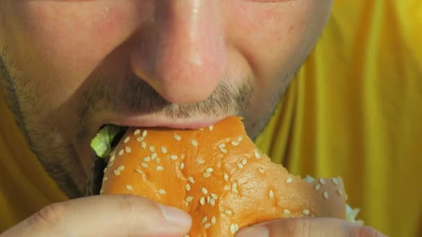 穿着黄色T恤的年轻人带着食欲吃汉堡包或汉堡包 嘴巴紧闭慢动作 咬和咀嚼垃圾食品 — 图库视频影像