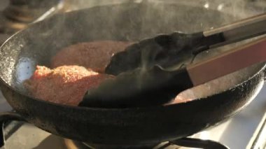 Tavada biftek kızartmak için büyük kırmızı bir parça biftek hazırlanıyor. Mutfak maşası bifteği yavaş çekimde çevirir. Yağ ve buhar damlaları havada uçuşuyor..